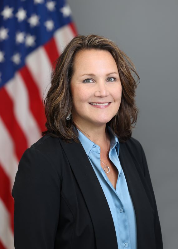 Lisa Gyorda with shoulder length brown, wavy hair in a blue colllared shirt and black blazer in front of the US flag
