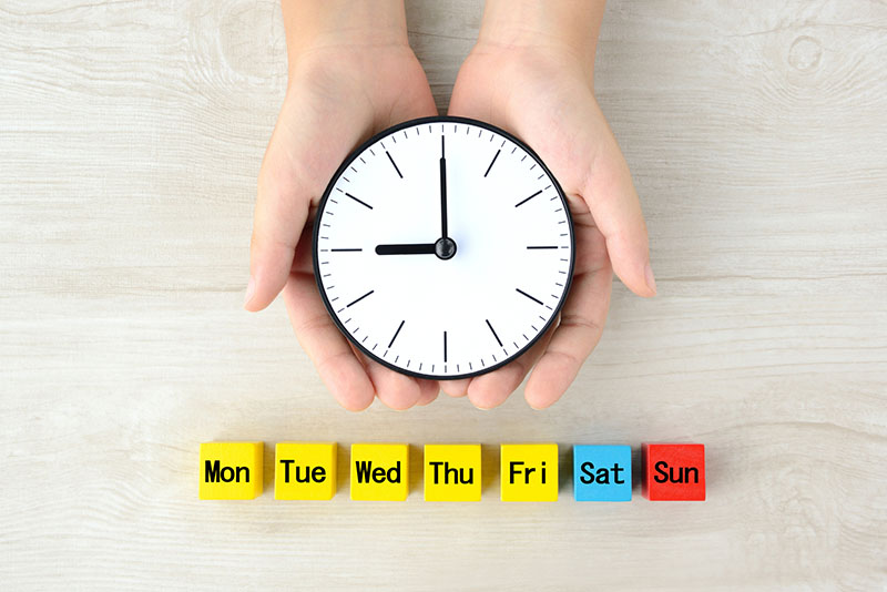 Abbreviated days of the week on blocks with hands holding a clock set to 9:00.