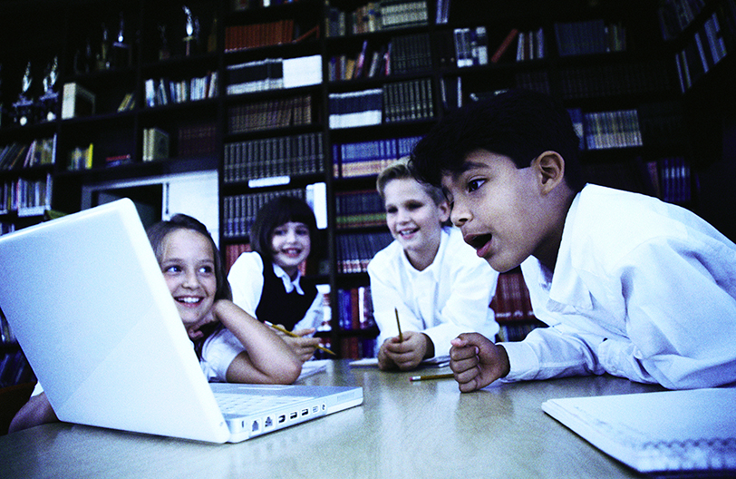 Elementary students at a library table looking at an open laptop computer.