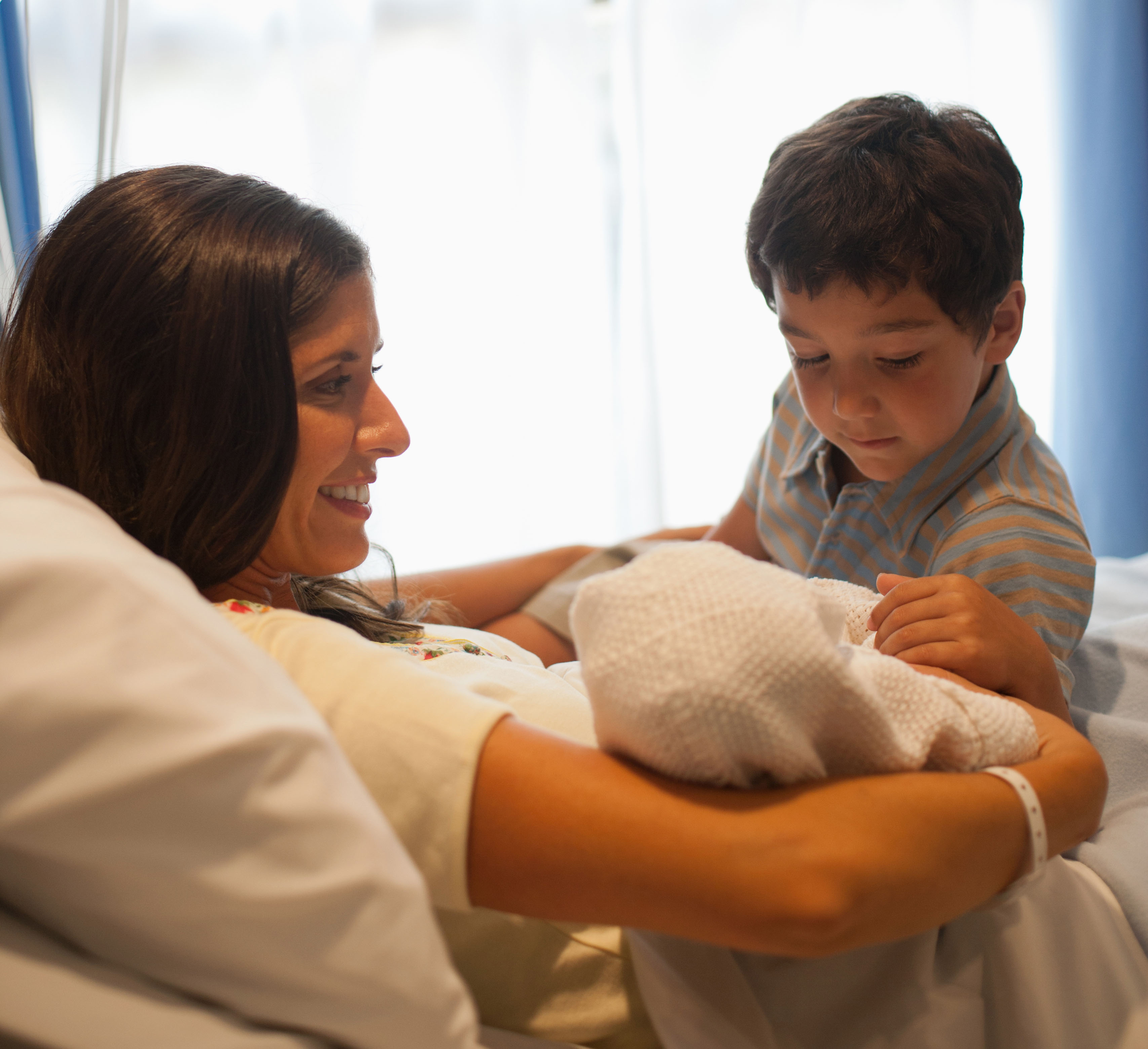 A mother holding her new baby with his big brother looking on